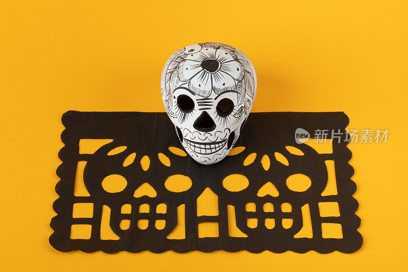 死亡的陶器头盖骨(calavera)在黑色papel picado在黄色背景，Dia de Muertos背景